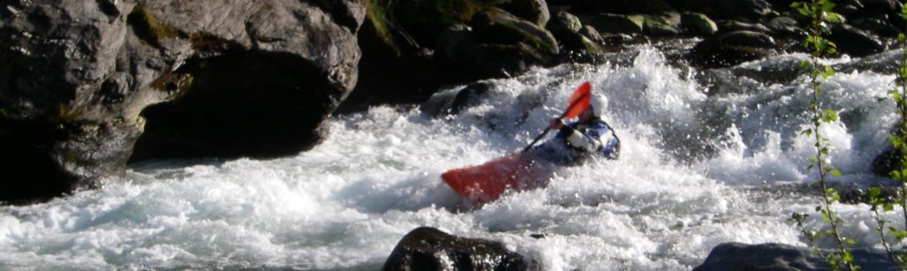 Eine Person in einem roten Kajak paddelt durch eine Stromschnelle in einem felsigen Fluss auf Korsika, der Insel der Schönheit. Wasser spritzt um sie herum, während sie durch die turbulente Strömung navigieren. Auf beiden Seiten des Flusses sind Felsen und Grünflächen zu sehen.