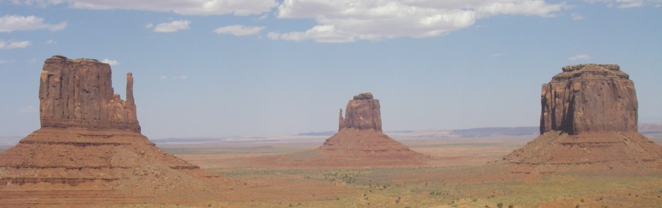 Ein Panoramablick auf das Monument Valley zeigt drei Sandsteinfelsen, die unter einem teilweise bewölkten Himmel aus der Wüstenlandschaft ragen.