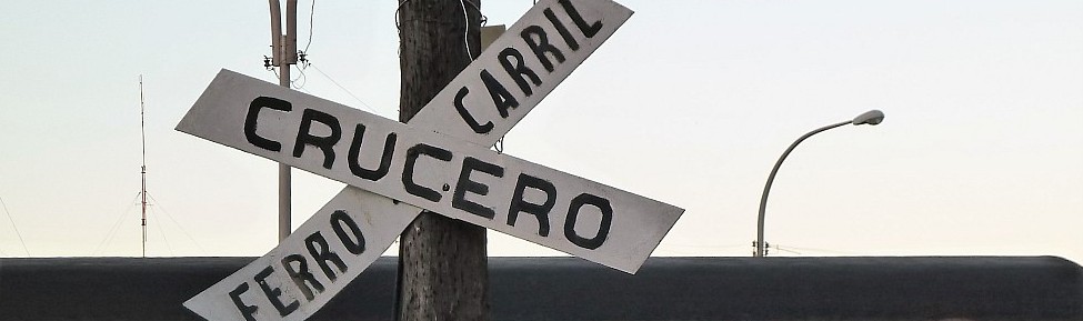 Ein hölzernes Bahnübergangsschild mit der Aufschrift „CRUCERO CARRIL FERRO“, angebracht an einem Strommast, vor einem klaren Himmel.
