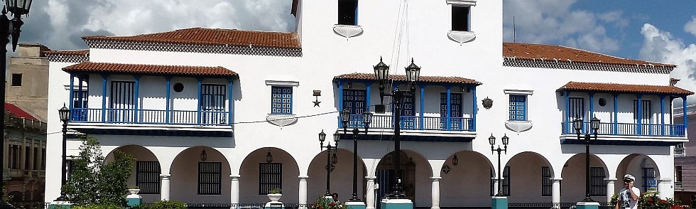 Ein weißes Gebäude im Kolonialstil mit blauen Verzierungen und Bogenfenstern, mit Balkonen und einem Uhrenturm, gelegen in einer sonnigen Gegend mit ein paar Straßenlaternen und Menschen davor.