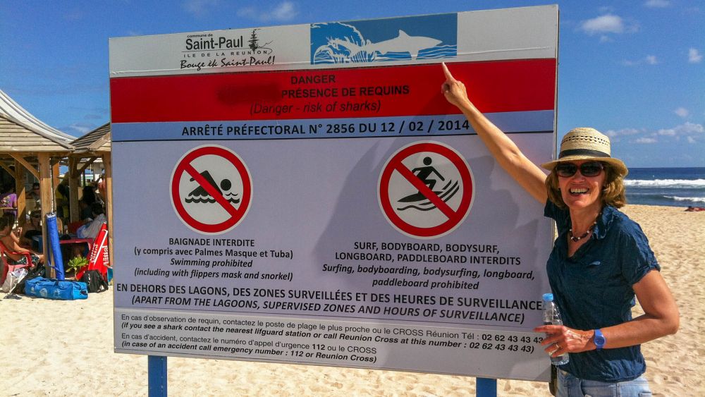 Gina zeigt auf ein Schild, das vor Haien im Meer warnt.
