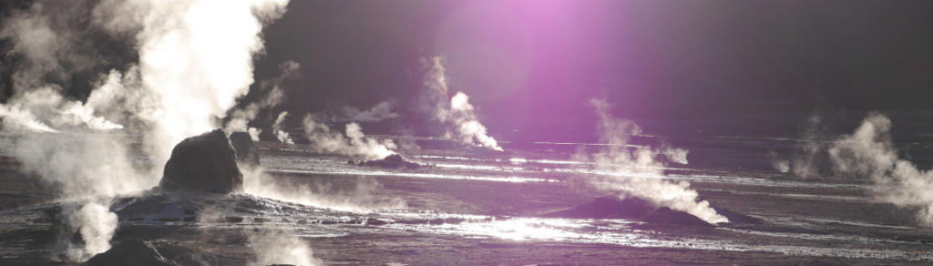 Dampf steigt aus mehreren Geysiren in einem geothermischen Feld auf, das von der Sonne von hinten beleuchtet wird und im oberen Teil des Bildes einen violetten Linsenreflex erzeugt. Die Landschaft ist überwiegend dunkel mit hellen Dampfformationen.