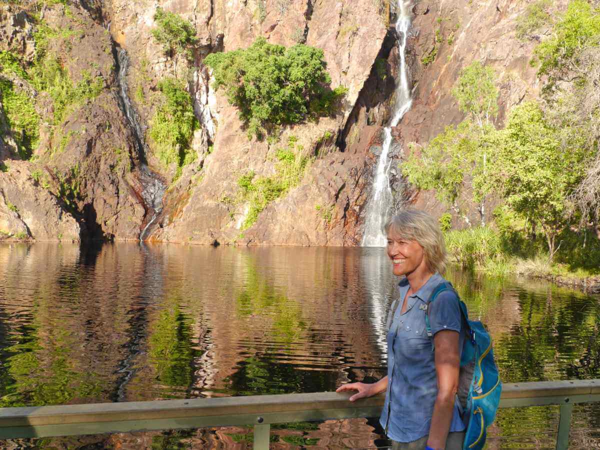 Gina steht vor einem kleinen See, im Hintergrund kommt ein schmaler Wasserfall die Felswand herunter.