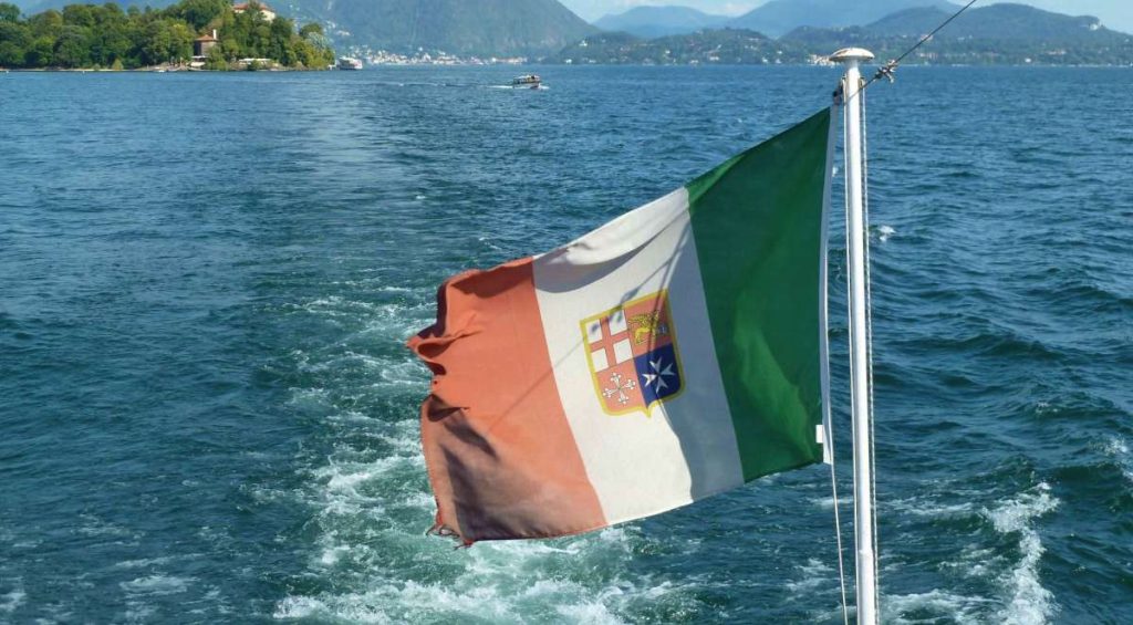 Eine dreifarbige Flagge mit vertikalen grünen, weißen und roten Streifen und einem Wappen in der Mitte weht auf dem Heck eines Bootes auf dem Bavenosee mit Bergen im Hintergrund.