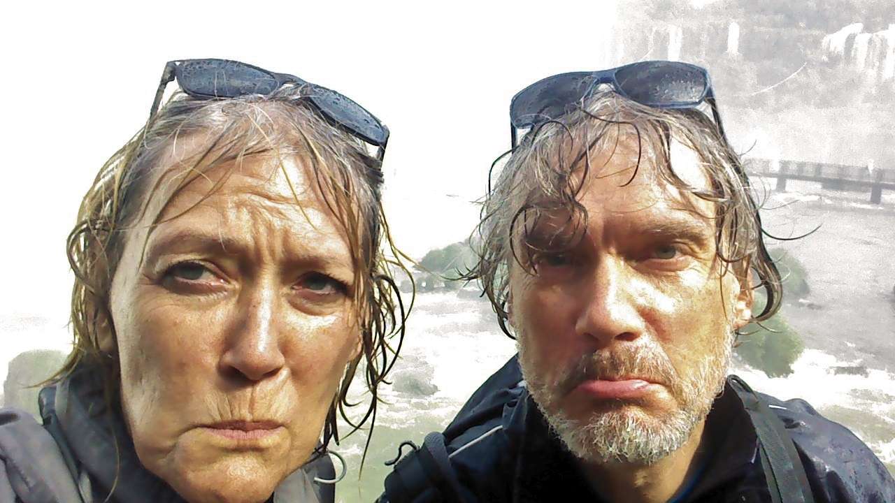 Zwei Personen mit nassen Haaren und Brillen auf dem Kopf stehen vor einem nebligen Wasserfall und zeigen beide einen übertrieben unglücklichen Gesichtsausdruck.