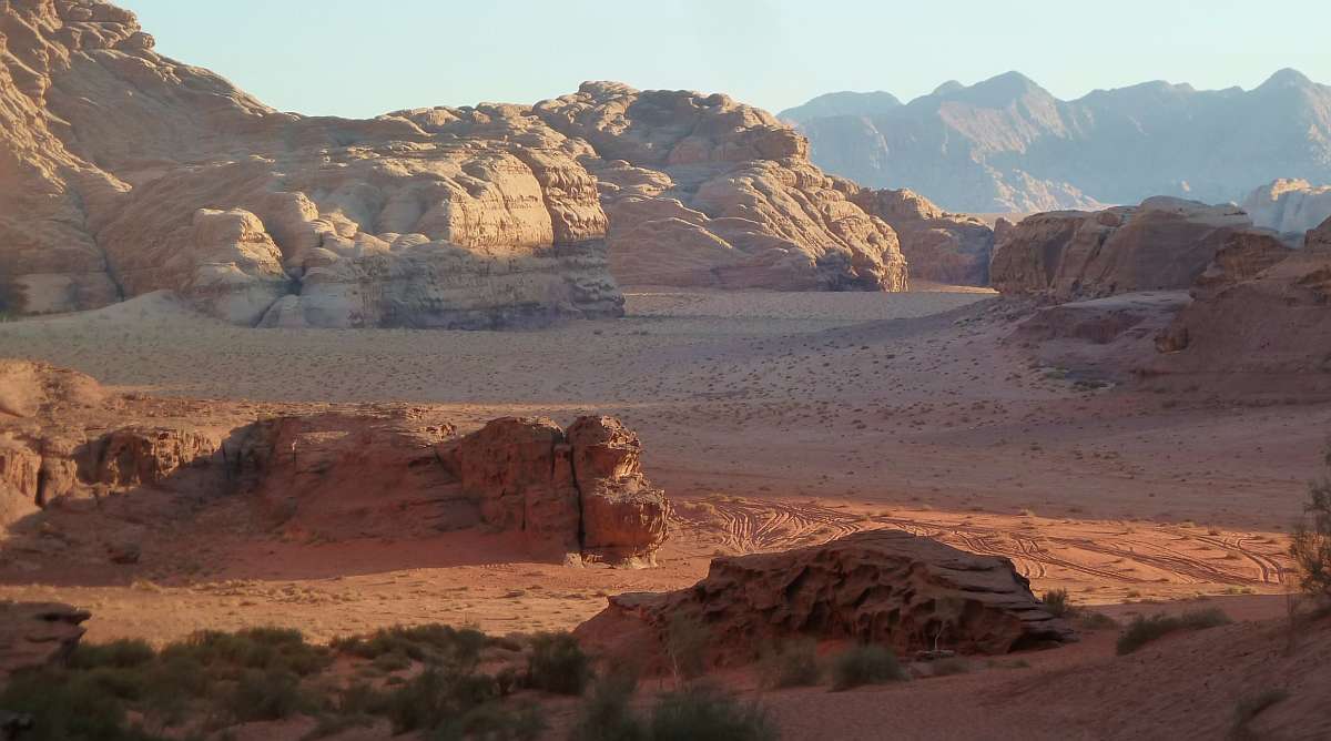 Wüstenlandschaft mit roten und beigen Felsformationen unter klarem Himmel, mit sandigem Gelände und verstreuter Vegetation im Vordergrund.