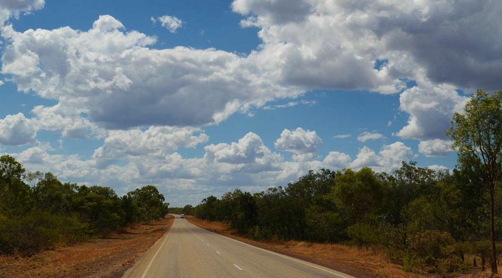 Unter einem teilweise bewölkten Himmel erstreckt sich in der Ferne eine gerade, asphaltierte Straße, auf beiden Seiten von Bäumen und trockener Vegetation gesäumt und bietet die perfekte Kulisse für einen abenteuerlichen Roadtrip.