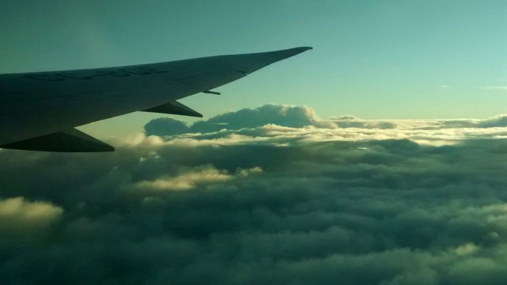 Blick aus einem Flugzeugfenster, der die Tragfläche vor dem Hintergrund dichter Wolken und eines sich aufhellenden Himmels zeigt.