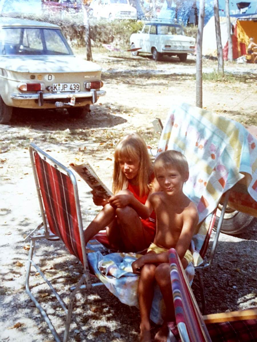 Zwei Kinder auf einem Liegstuhl, im Hintergrund das Auto.