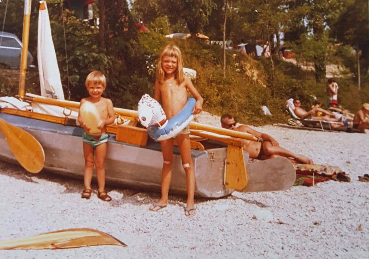 Zwei Kinder mit Schwimmringen stehen vor einem Boot am Strand.