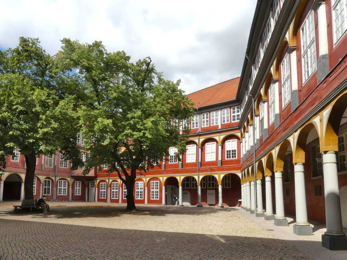 Hof mit weißen Säulengängen, roter Fassade und großem Baum.