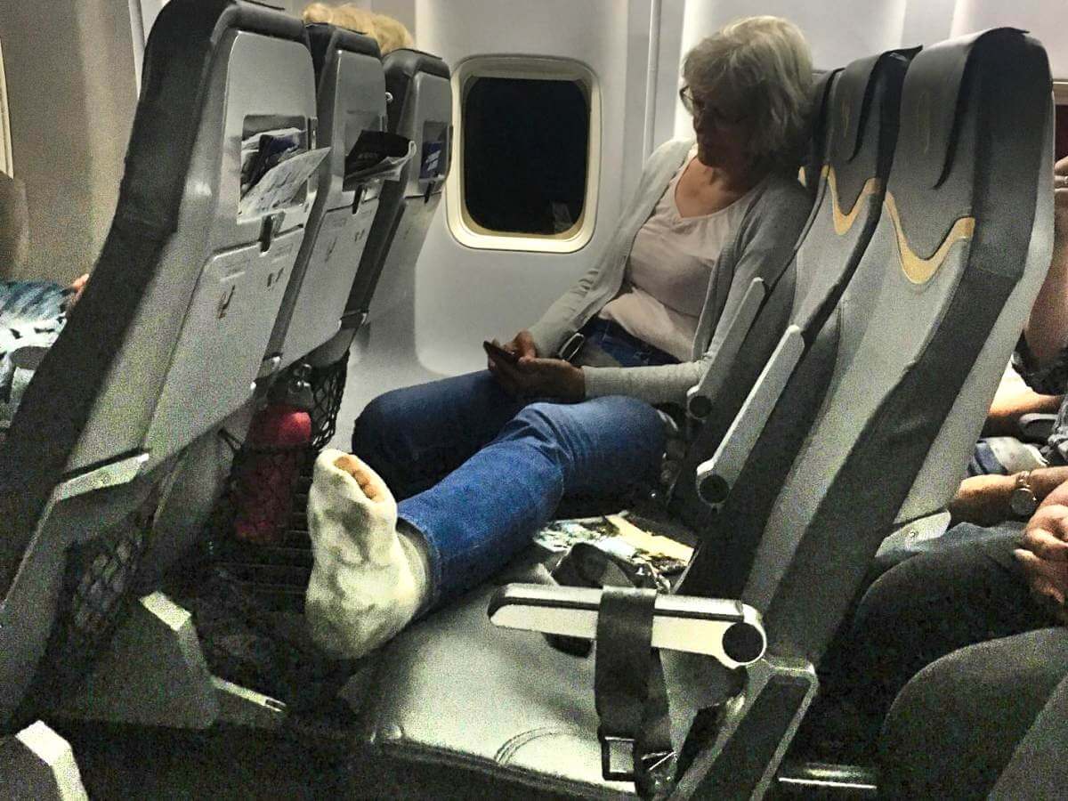 Gina mit ausgestrecktem Bein in Sitzreihe im Flugzeug.