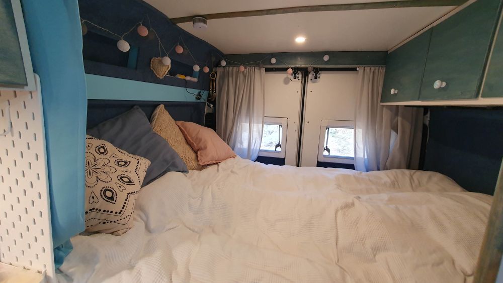 Bett im Camper mit weißer Überdecke und Kissen in hellen Farben.