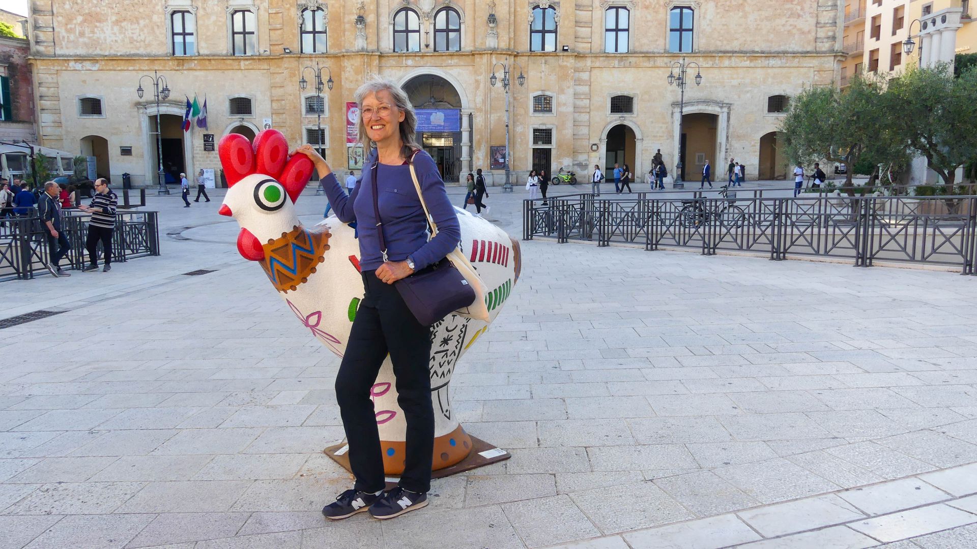 Gina steht neben einer bunten Skulptur eines Hahns.