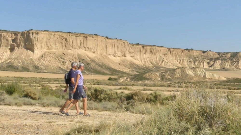 Gina und Marcus laufen auf einem Pfad durch die Wüste, im Hintergrund eine ockerfarbene Steilwand.