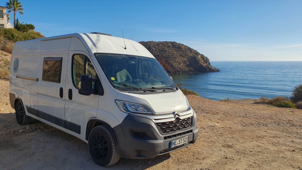 Wohnmobil steht an der Küste in Spanien, vor blauem Meer und Felsenkap.