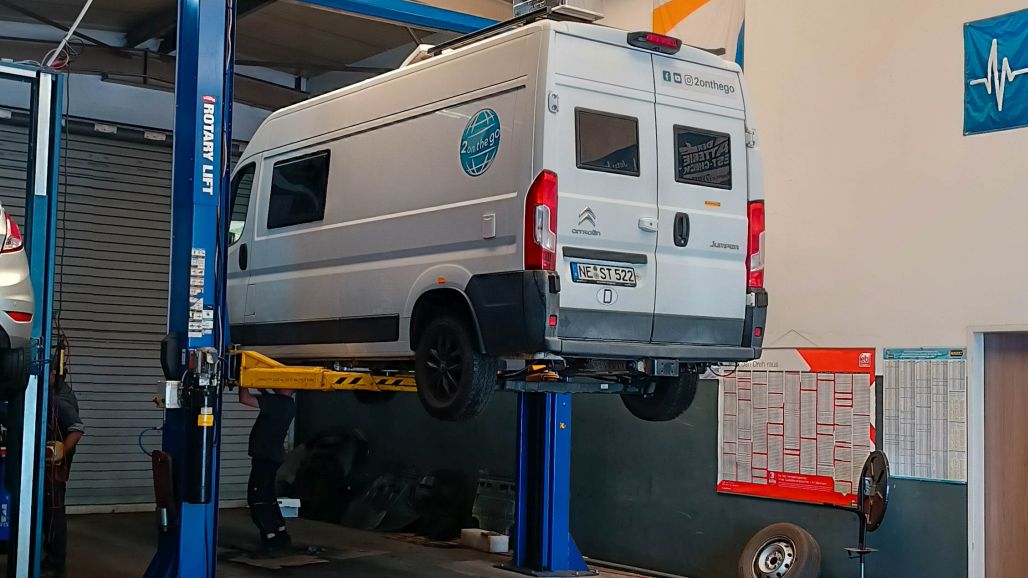 Weißer Kastenwagen auf einer hydraulischen Hebebühne in einer Garage, darunter arbeitet ein Mechaniker.
