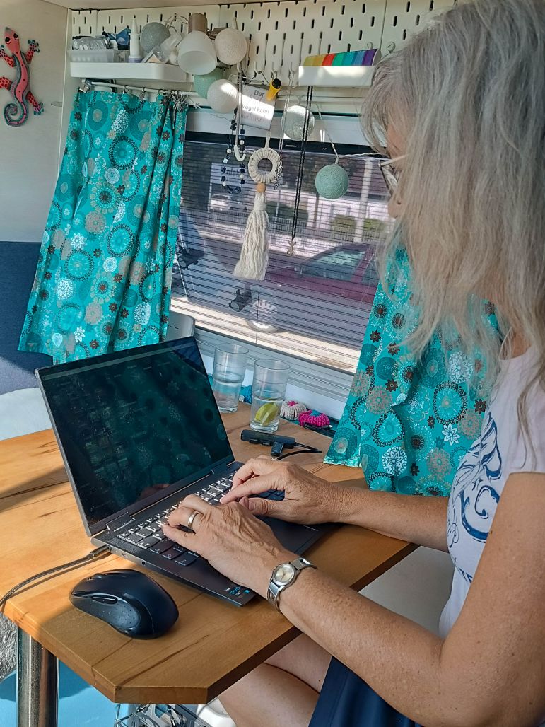 Eine Person mit grauem Haar tippt an einem Laptop an einem kleinen Holztisch im Van mit türkisfarbenen Vorhängen und Dekorationsgegenständen.