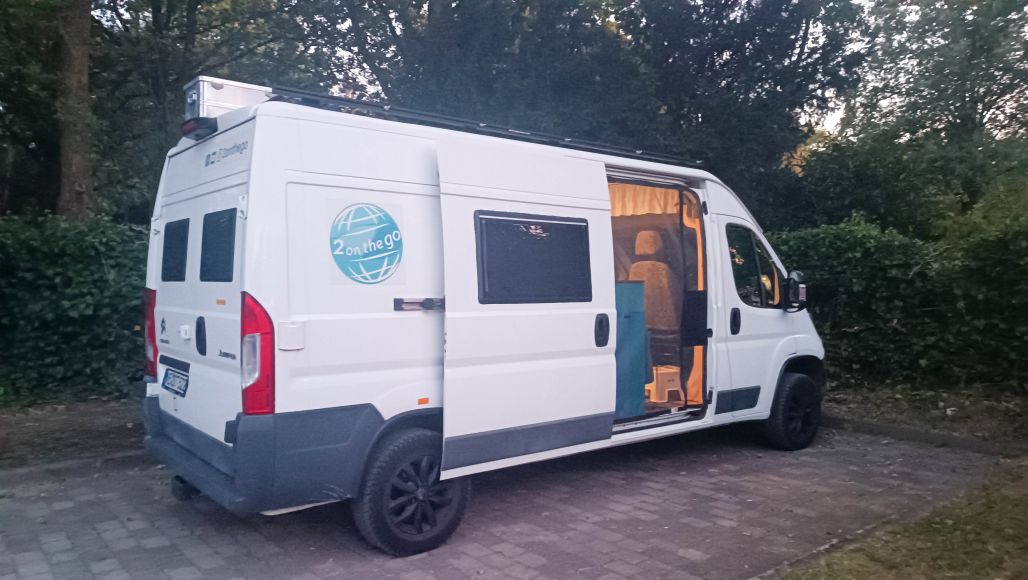 Weißer Campervan, der auf einer geschotterten Fläche geparkt ist, umgeben von Grün. Die Schiebetür des Vans ist geöffnet und an der Seite befindet sich ein Logo mit dem Text „2 on the go“.