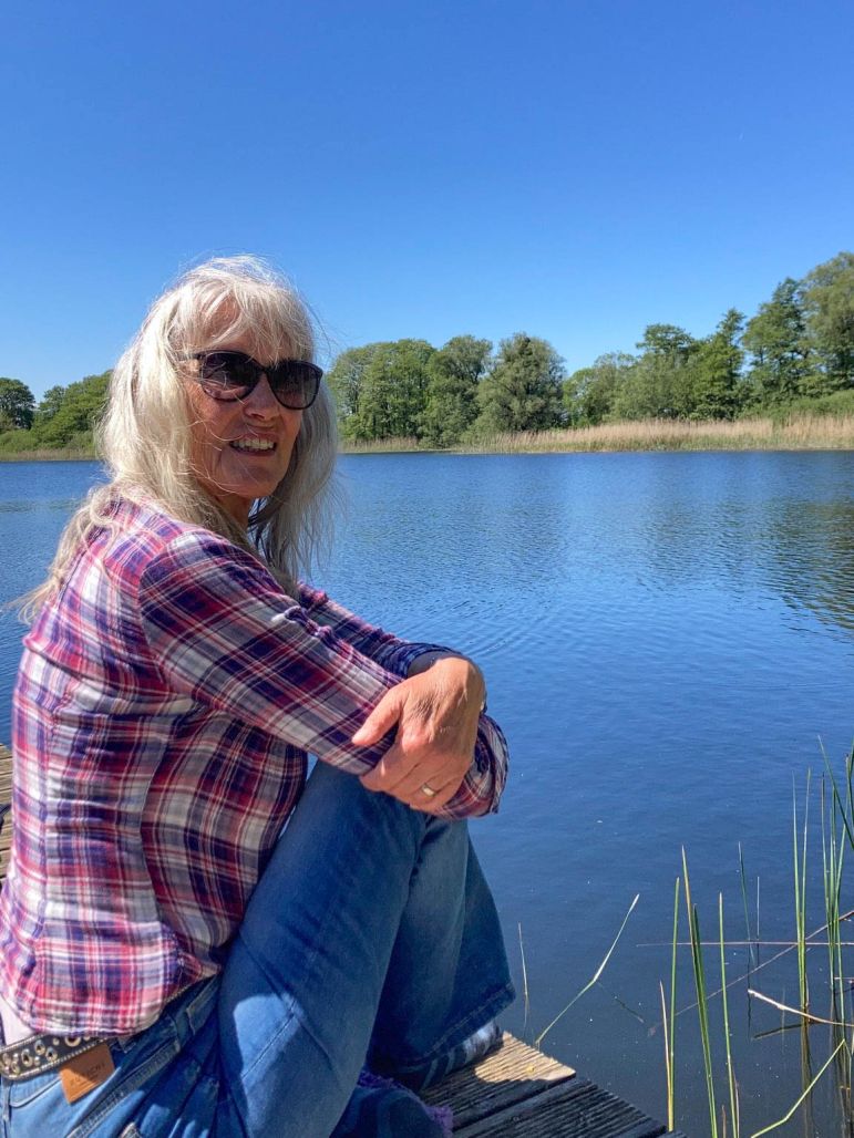Gina in kariertem Hemd und Sonnenbrille sitzt an einem ruhigen See unter einem klaren blauen Himmel.