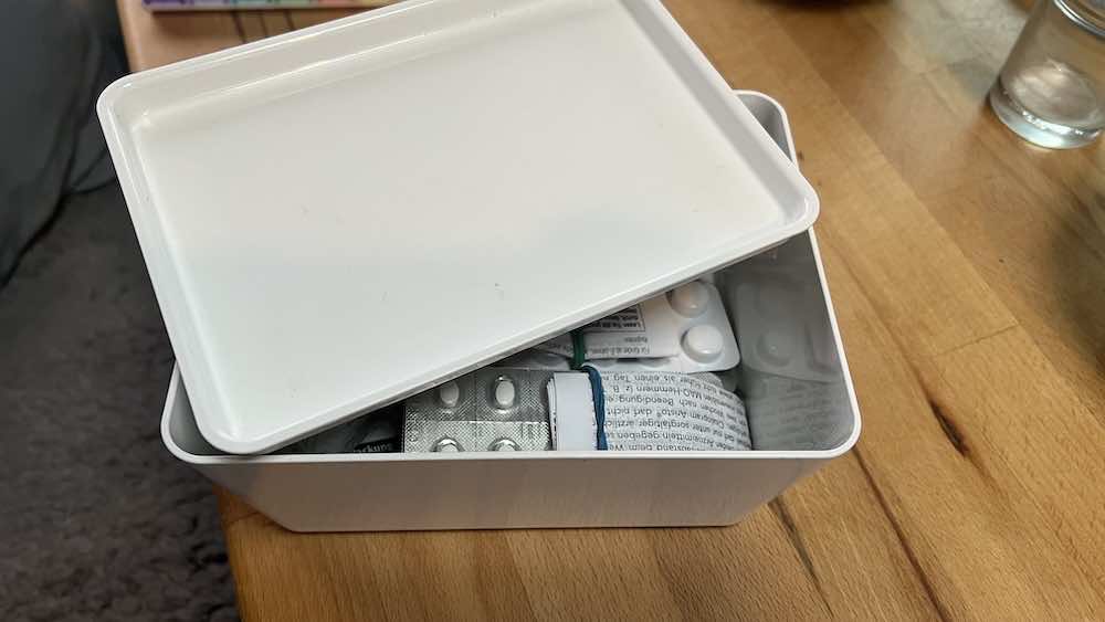 Eine weiße Kunststoffbox mit teilweise geöffnetem Deckel, die verschiedene Blisterpackungen mit Medikamenten und Pillenpackungen auf einem Holztisch enthält.