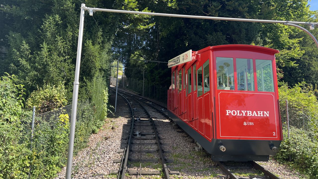 Die rote Polybahn fährt an einem sonnigen Tag auf einer eingleisigen Strecke durch eine grüne, baumbestandene Gegend bergauf.
