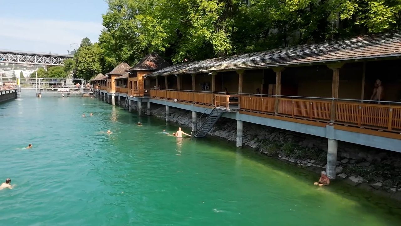 Menschen schwimmen in einem türkisfarbenen Fluss neben einem hölzernen Ufergebäude mit einer schattigen Terrasse, umgeben von grünen Bäumen und einer Brücke im Hintergrund.