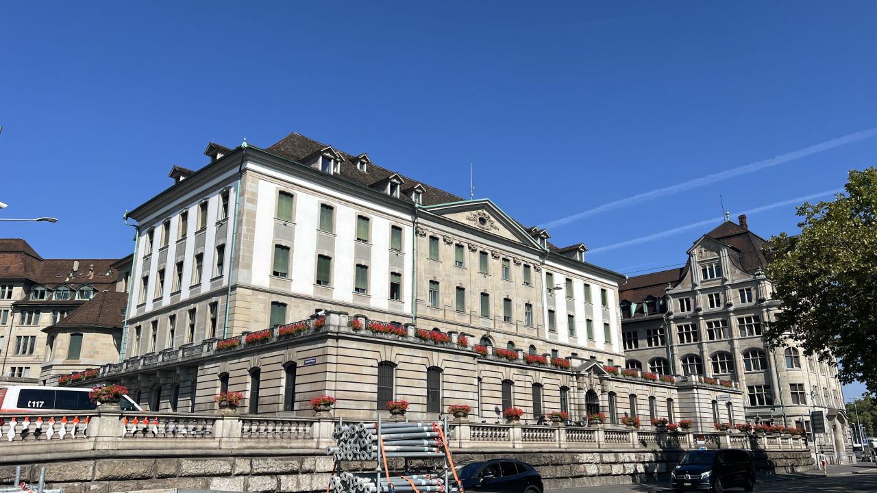 Ein großes, historisches europäisches Gebäude mit Fensterreihen und roten Blumen vor einem strahlend blauen Himmel, mit anderen Gebäuden und geparkten Autos in der Nähe.