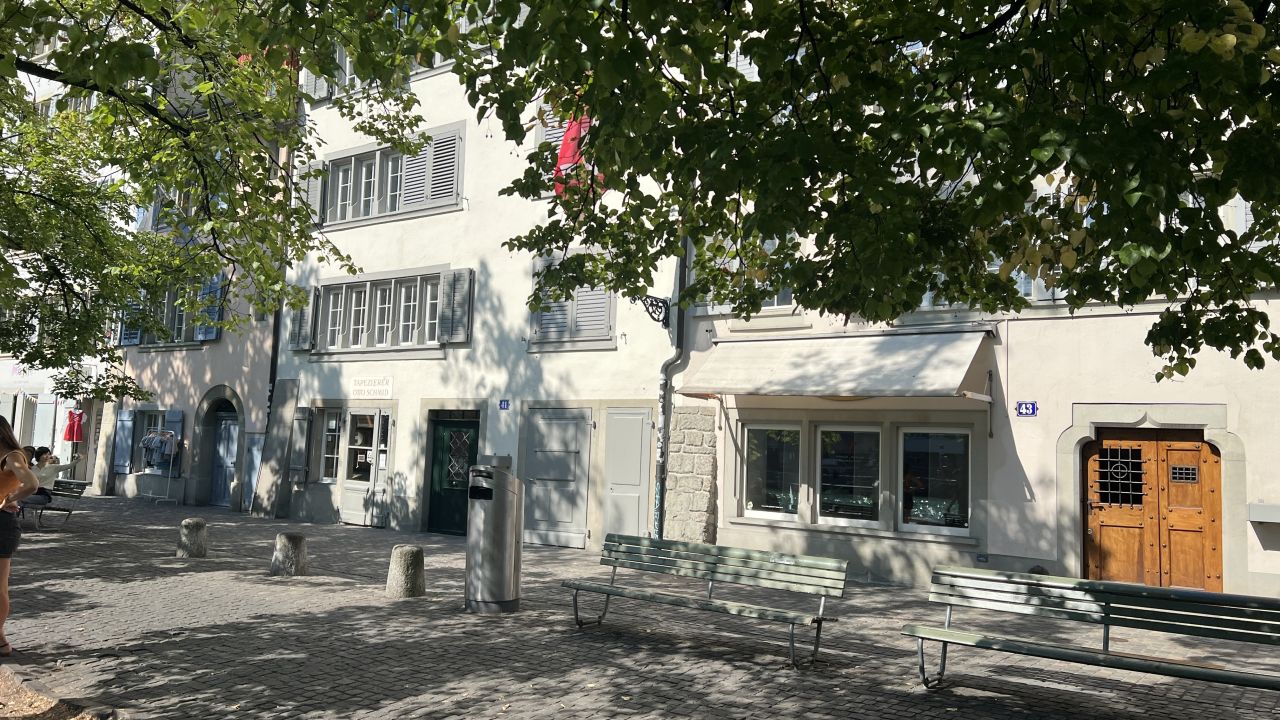 Eine sonnenbeschienene Straße mit Bänken, Bäumen und mehreren weißen Gebäuden mit verschlossenen Fenstern und einer Holztür. Die Schatten des Laubes fallen auf das Kopfsteinpflaster.