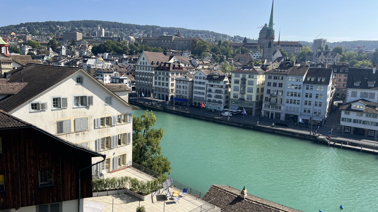 Ansicht von Zürich mit traditionellen Gebäuden entlang der türkisfarbenen Limmat, einer Kirche mit grünem Turm und Hügeln im Hintergrund unter einem klaren Himmel.