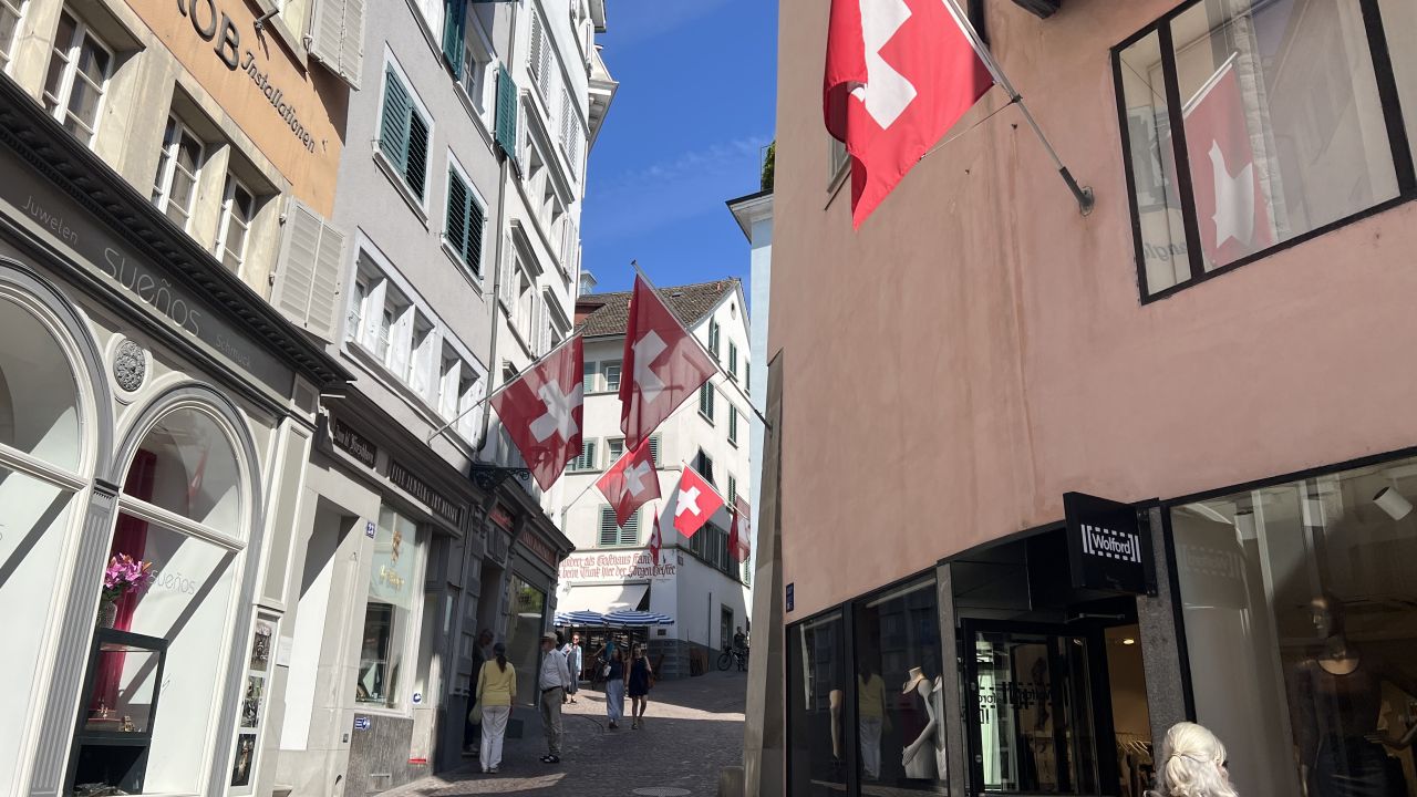 Eine schmale Kopfsteinpflasterstraße mit Geschäften und vielen Schweizer Fahnen an den Häusern unter einem strahlend blauen Himmel.
