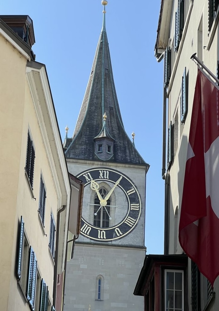 Ein großer Uhrenturm mit römischen Ziffern steht an einem sonnigen Tag zwischen Gebäuden; eine Schweizer Flagge hängt am rechten Bildrand.