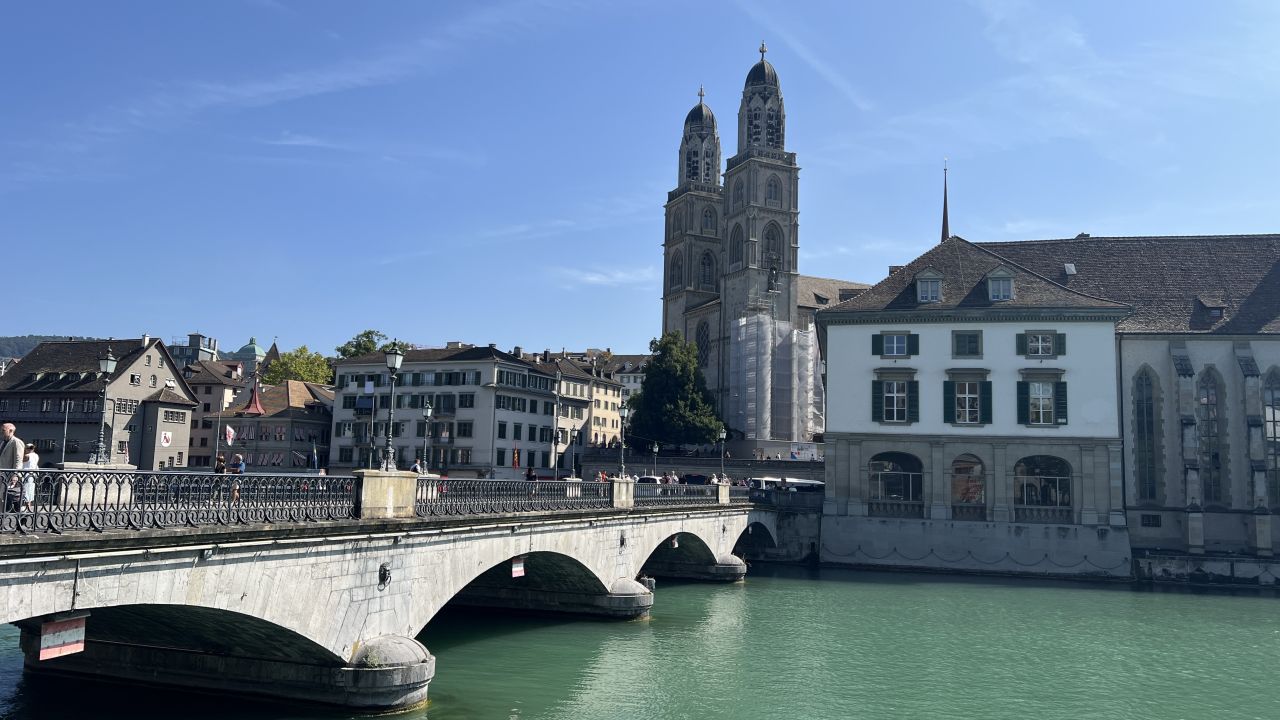 Steinbrücke über die türkisfarbene Limmat mit historischen Gebäuden und den Zwillingstürmen der Grossmünsterkirche unter einem klaren blauen Himmel in Zürich, Schweiz.