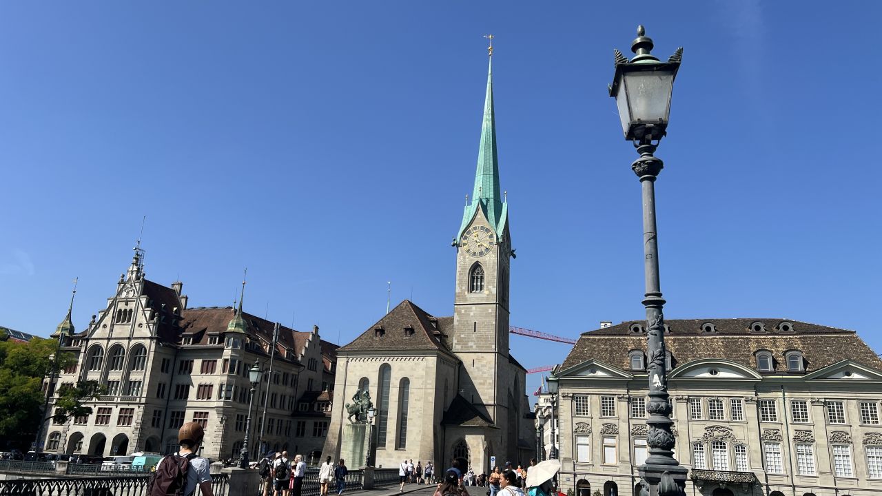 Menschen gehen auf einer Brücke in Richtung der Kirche Fraumünster mit einem hohen grünen Turm, umgeben von historischen Gebäuden unter einem klaren blauen Himmel. Im Vordergrund steht eine Straßenlaterne.