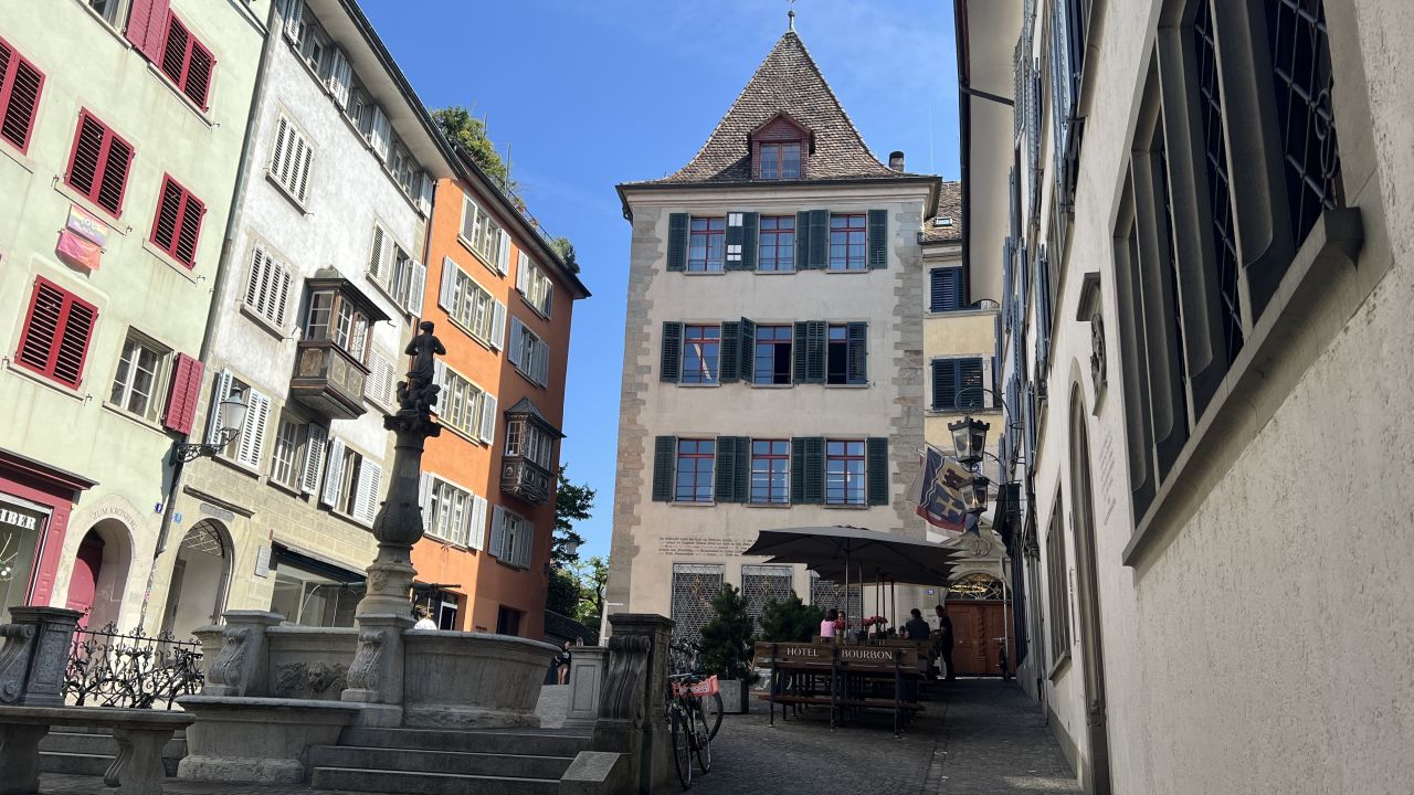 Eine schmale Kopfsteinpflasterstraße mit bunten Gebäuden, einem steinernen Brunnen im Vordergrund und einem Straßencafé, in dem Menschen unter Sonnenschirmen sitzen.