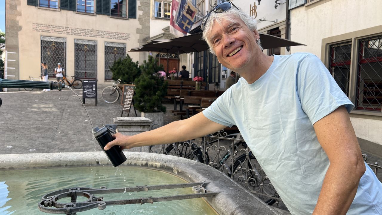 Ein Mann in einem hellblauen Hemd lächelt, während er eine schwarze Flasche an einem öffentlichen steinernen Wasserbrunnen in Zürich füllt.