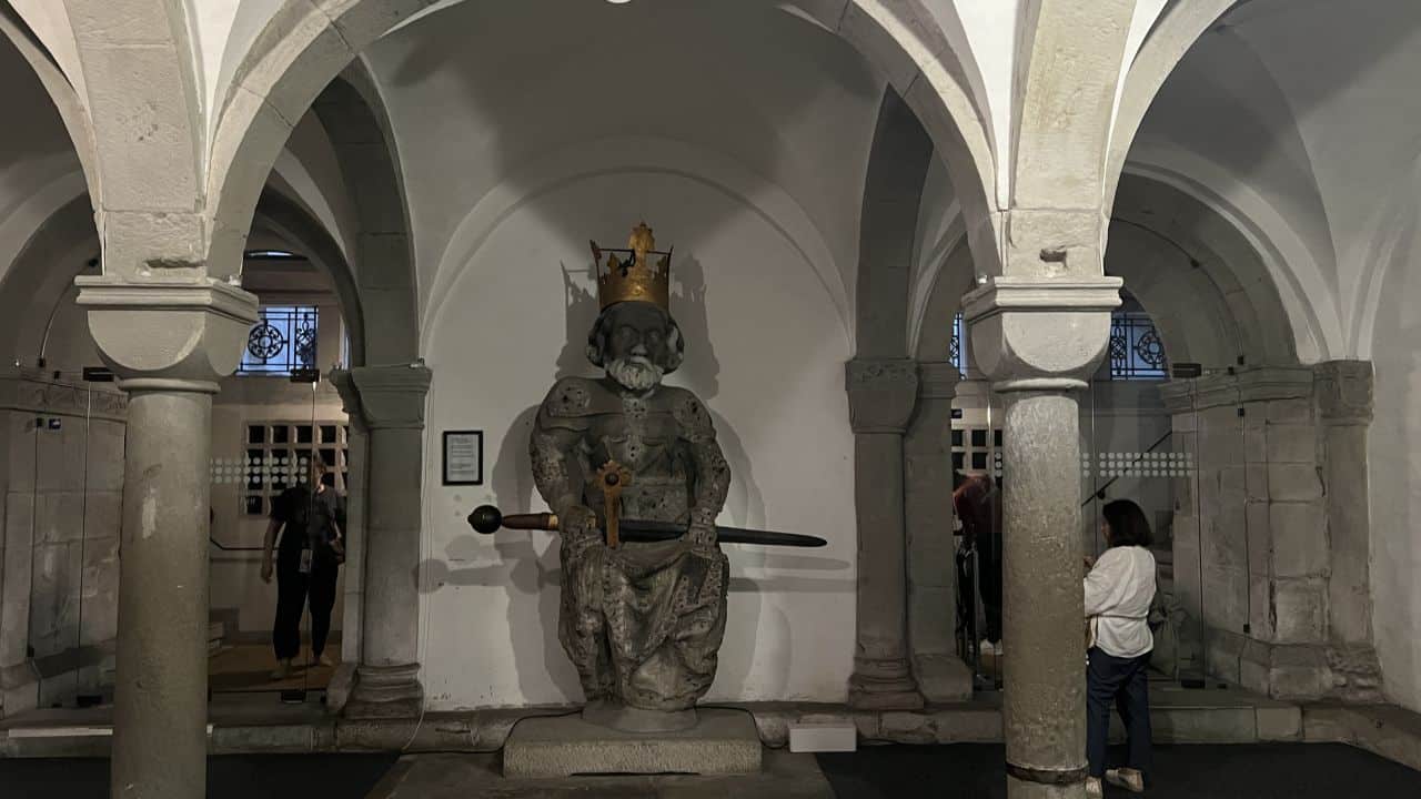 Eine steinerne Statue einer gekrönten Figur mit einem Schwert steht in einem gewölbten Raum; mehrere Personen stehen in der Nähe und beobachten das Exponat.