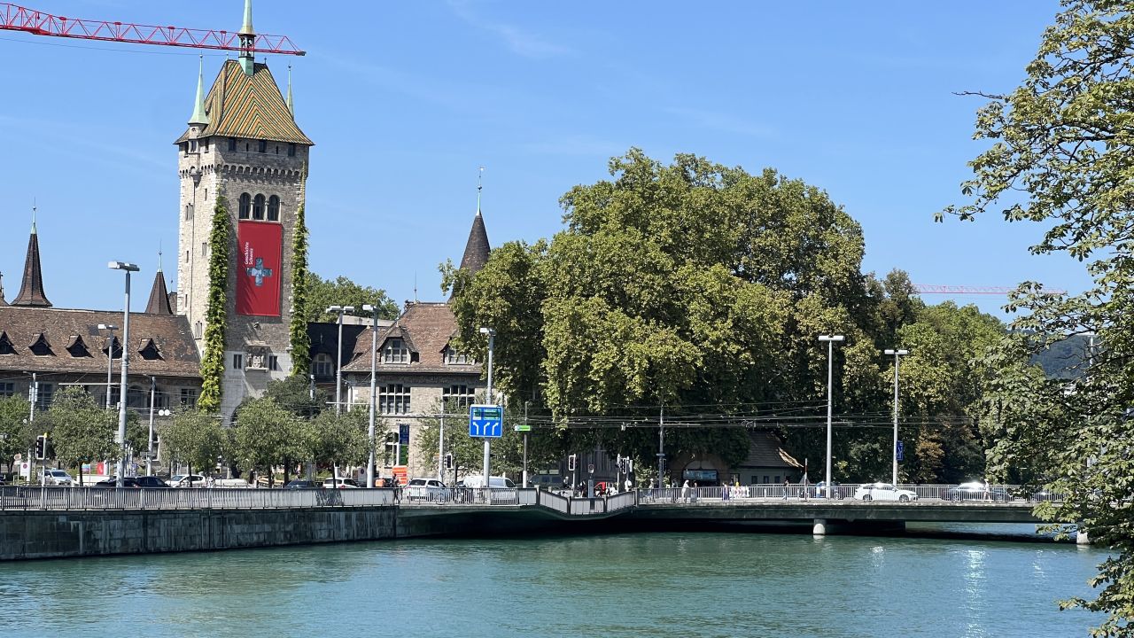 Ein Fluss fließt unter einer Brücke in der Nähe eines historischen Steingebäudes mit Uhrenturm und roter Fahne, umgeben von Bäumen und einem klaren blauen Himmel.