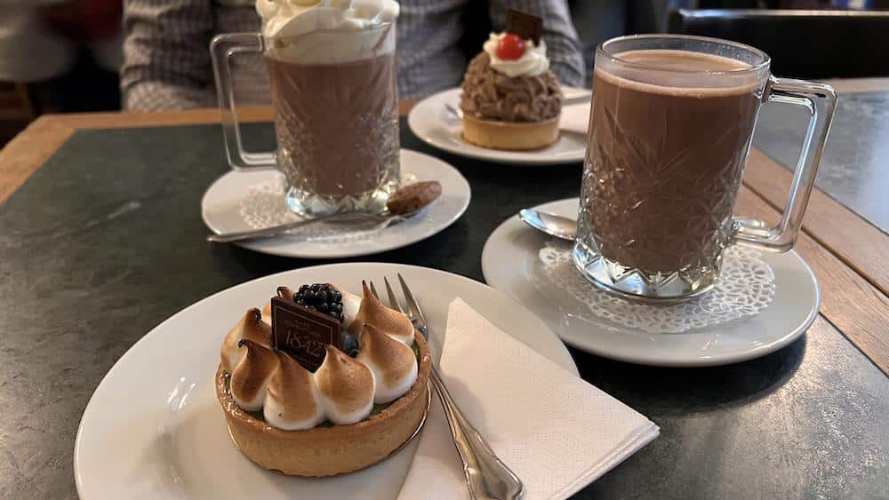 Zwei Becher mit heißer Schokolade und zwei einzelne Desserts werden auf weißen Tellern auf einem dunklen Tisch serviert, daneben liegen Utensilien und Servietten.