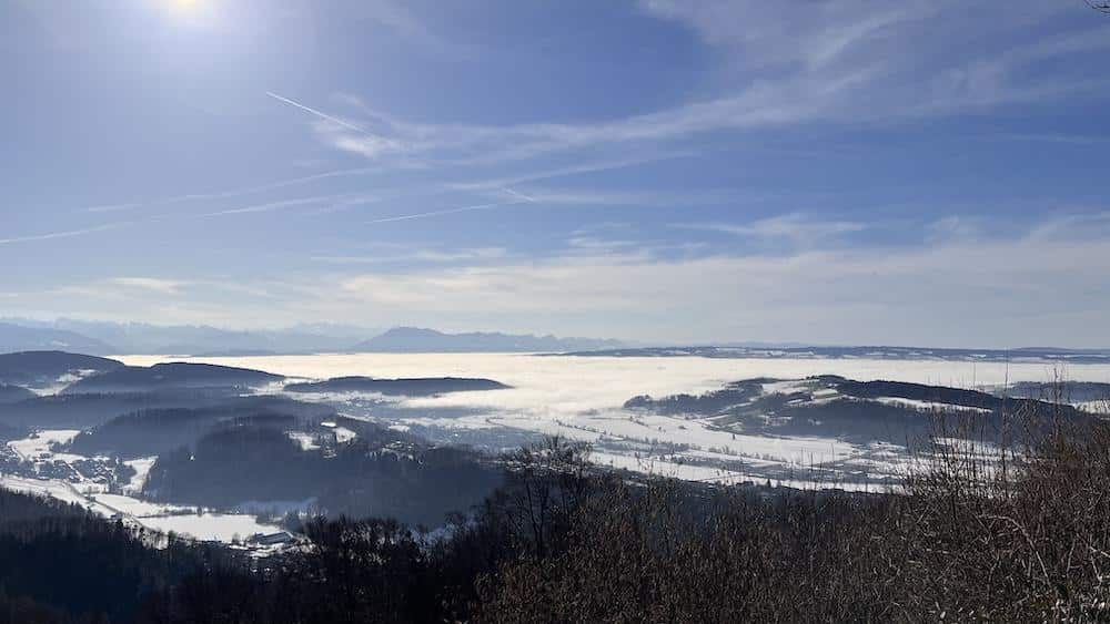 Eine Landschaftsansicht eines nebelverhangenen Tals mit umliegenden Hügeln, Schneeflecken, entfernten Bergen und einer hellen Sonne an einem teilweise bewölkten Himmel.