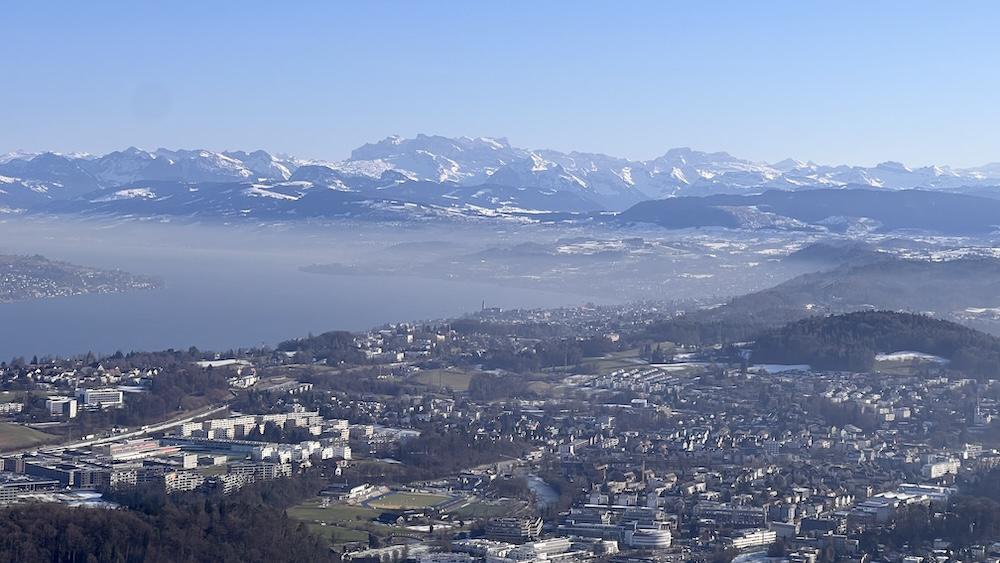 Luftaufnahme von Zürich mit Gebäuden, einem See und entfernten schneebedeckten Bergen unter einem klaren blauen Himmel.