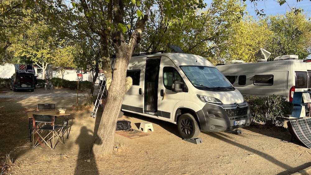 Ein weißes Wohnmobil ist auf einem Campingplatz neben einem Baum geparkt, mit Campingstühlen und einem Tisch in der Nähe. Ein weiteres Wohnmobil und Bäume sind im Hintergrund zu sehen.