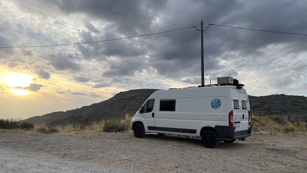 Ein weißes Wohnmobil ist auf einer Schotterstraße in der Nähe einer Berglandschaft bei Sonnenuntergang geparkt, mit bewölktem Himmel und einem Strommast im Hintergrund.