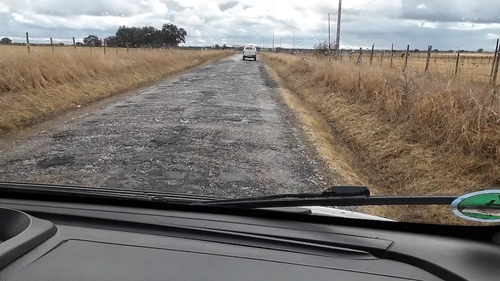 Blick aus dem Inneren eines Wohnmobils, das auf einer schmalen, abgenutzten Landstraße mit trockenem Gras auf beiden Seiten und einem Auto vor ihm bei bewölktem Himmel fährt.