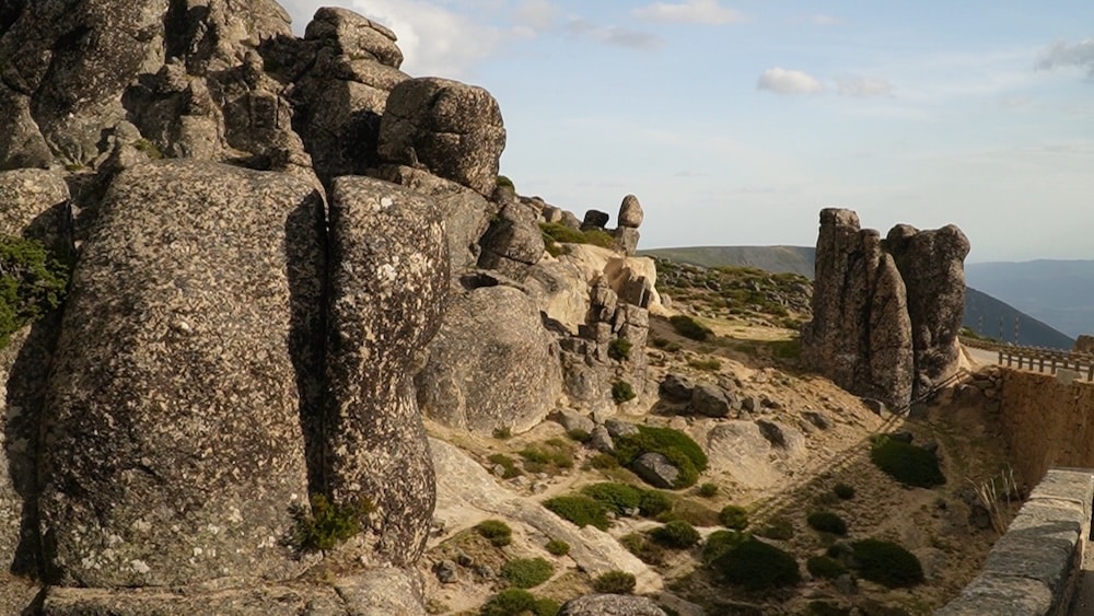 Große verwitterte Felsformationen und verstreute Felsbrocken auf einem trockenen, felsigen Hang unter einem teilweise bewölkten Himmel. Eine Steinmauer verläuft entlang der rechten Seite der Szene.