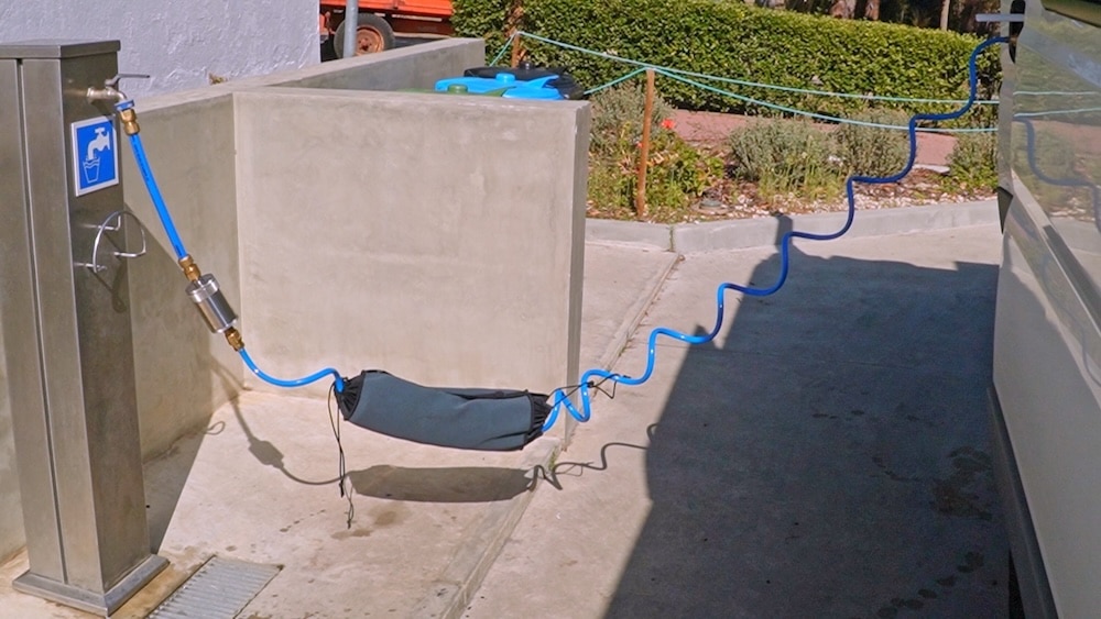 Ein blauer Schlauch verbindet eine Wasserzapfstelle mit einem Wohnmobil. In der Mitte hängt eine schwarze Tasche, die den Schlauch über dem Boden hält.