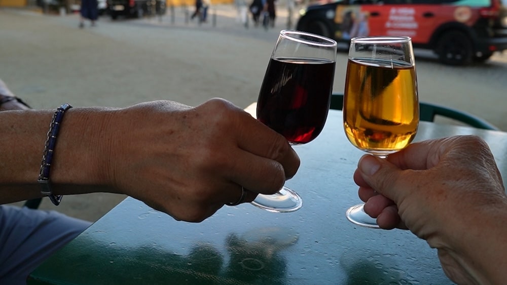 Zwei Personen stoßen an einem grünen Tisch im Freien mit Gläsern mit rotem und bernsteinfarbenem Portwein an, im Hintergrund sind unscharfe Menschen und ein Auto zu sehen.
