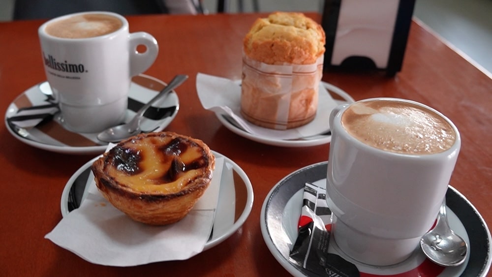 Zwei Tassen Kaffee, ein Pastel de Nata und ein Muffin stehen auf Untertassen mit Löffeln und Servietten auf einem braunen Tisch.
