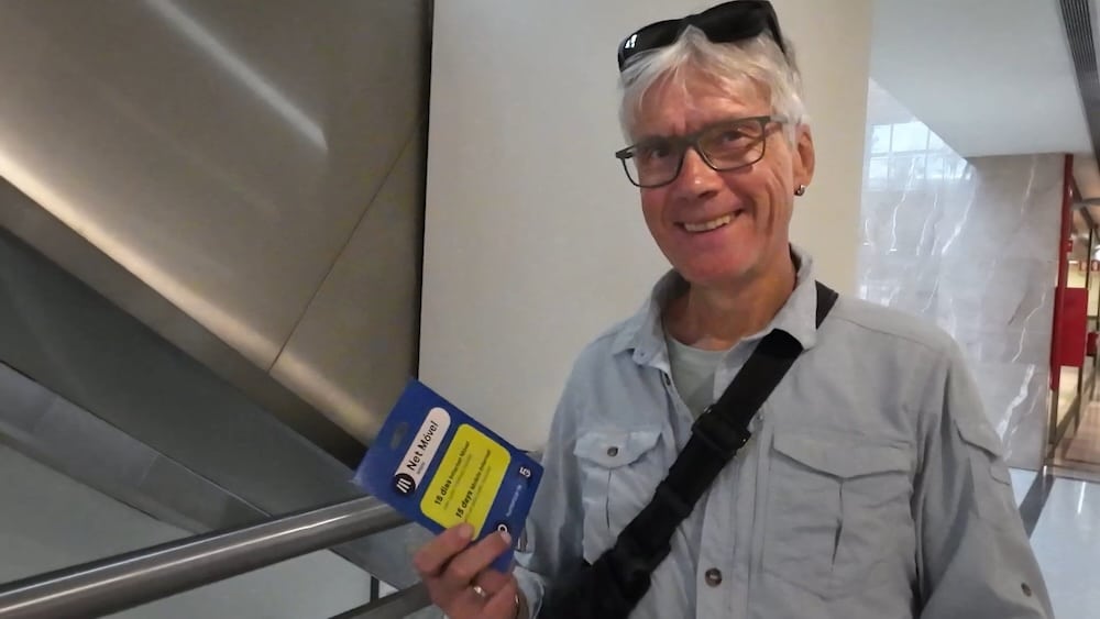 Ein Mann mit grauem Haar und Brille, hellem Hemd und Umhängetasche, steht in einem Haus, hält eine blaue SIM-Kartenpackung in der Hand und lächelt in die Kamera.