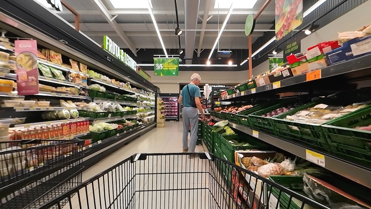 Ein Gang in einem Lebensmittelgeschäft mit Regalen mit Produkten und verpackten Lebensmitteln; eine Person steht und wählt Artikel aus, von einem Einkaufswagen aus gesehen.